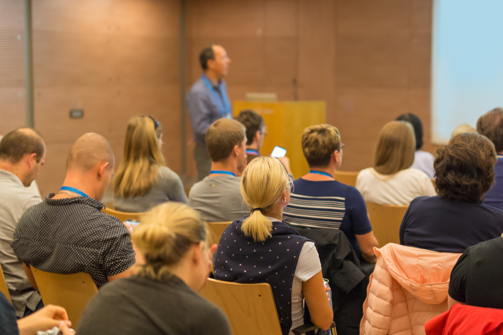 A male lecturer stands at the front of a group of seated people. The group are watching a presentation screen..