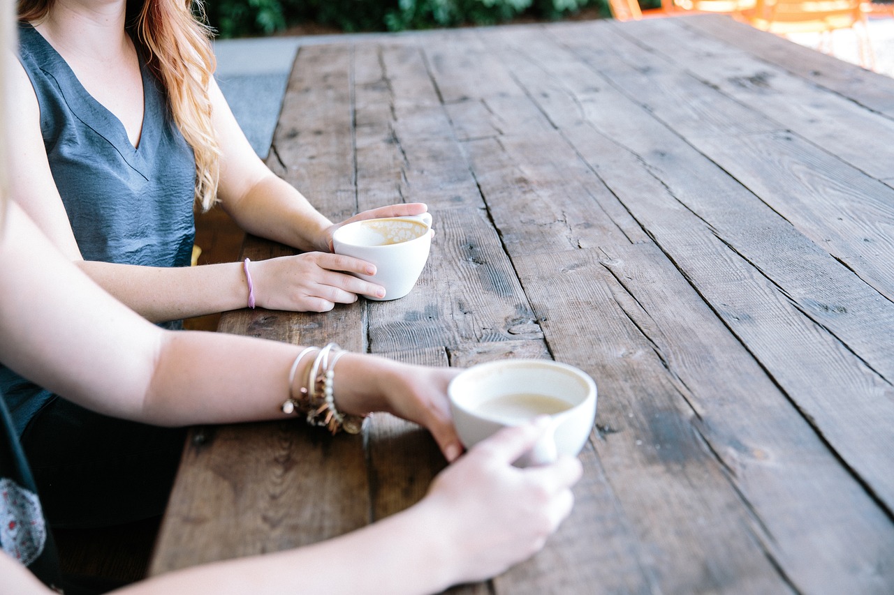 Two people sit at a wooden picnic bench with cups of coffee