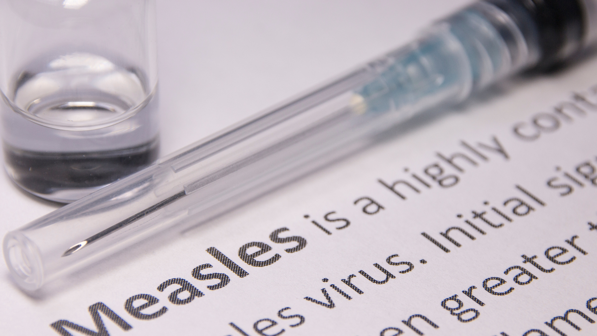 The image shows a document with the word measles and an explanation that the virus is highly contagious. The image also shows a vaccine vial and needle.