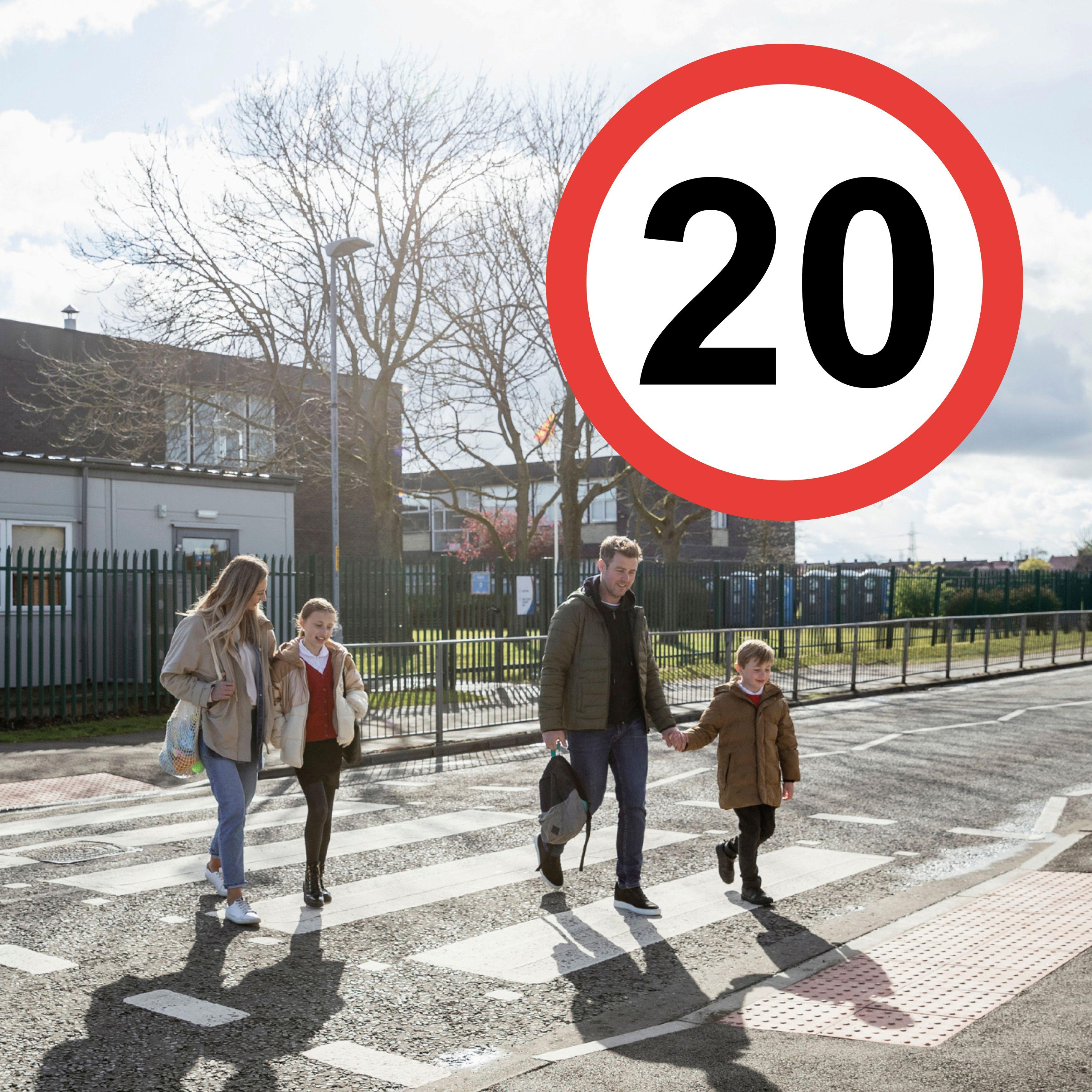 Two adults and two children crossing the road. A 20mph symbol has been added to the top right hand corner of the image