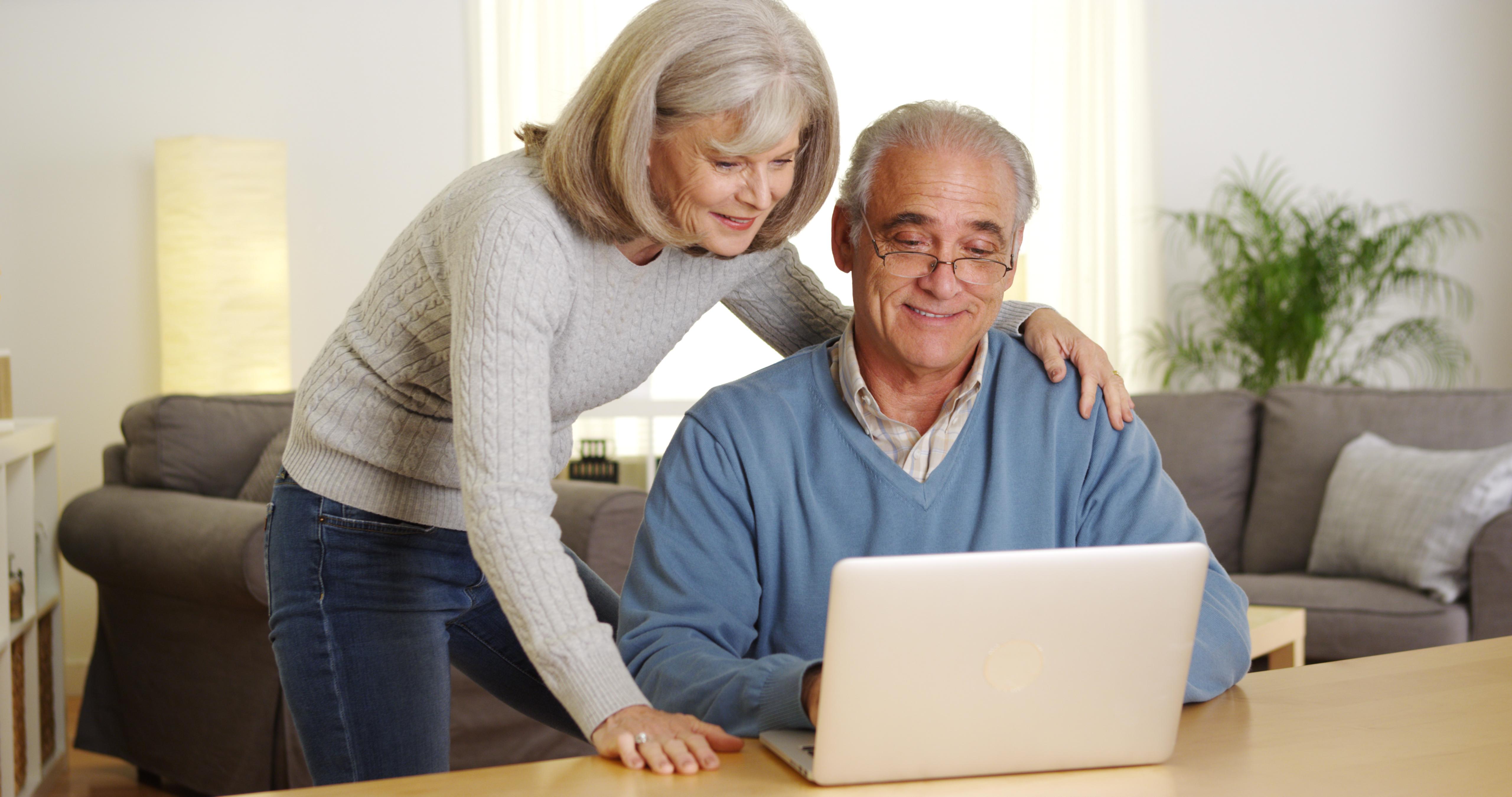 One older man and one older woman looking at a laptop, the man is sat the woman is leaning over him, they are both engaged by what is on the screen.