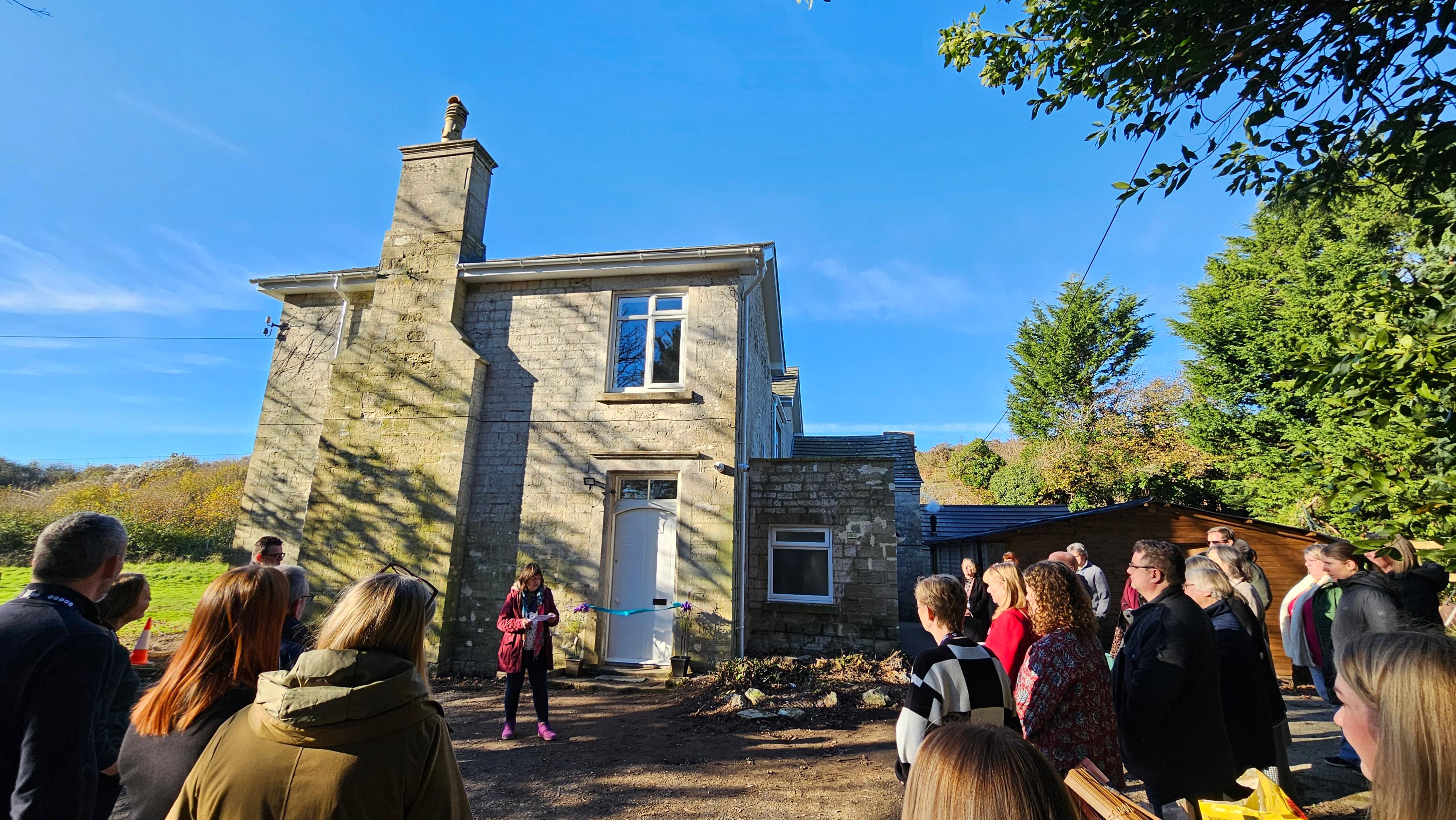 A group of people gather outside a Victorian farmhouse to witness the opening of a new housing scheme