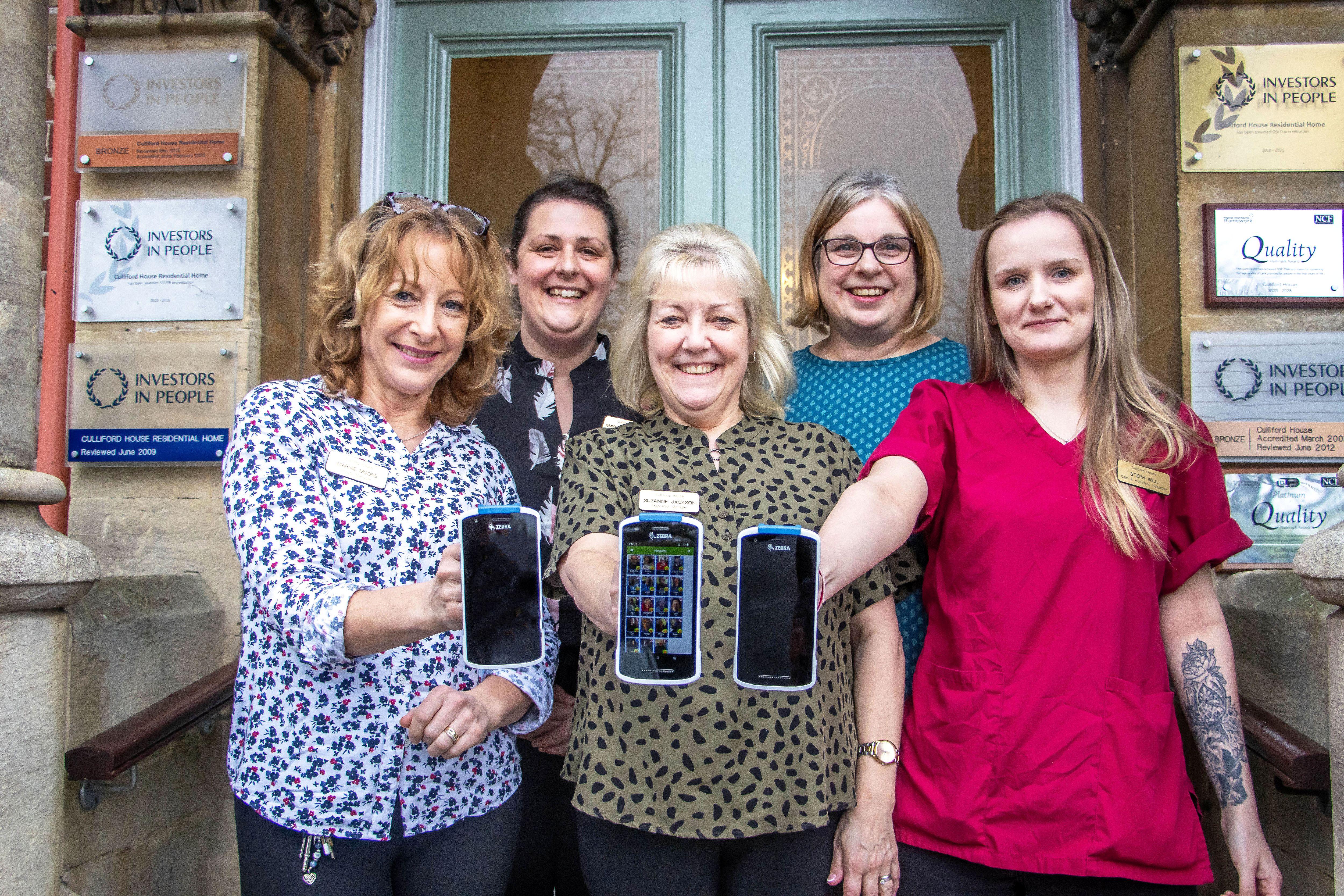 Five women are standing in front of a door entrance. The three women in the front are holding out digital handsets.