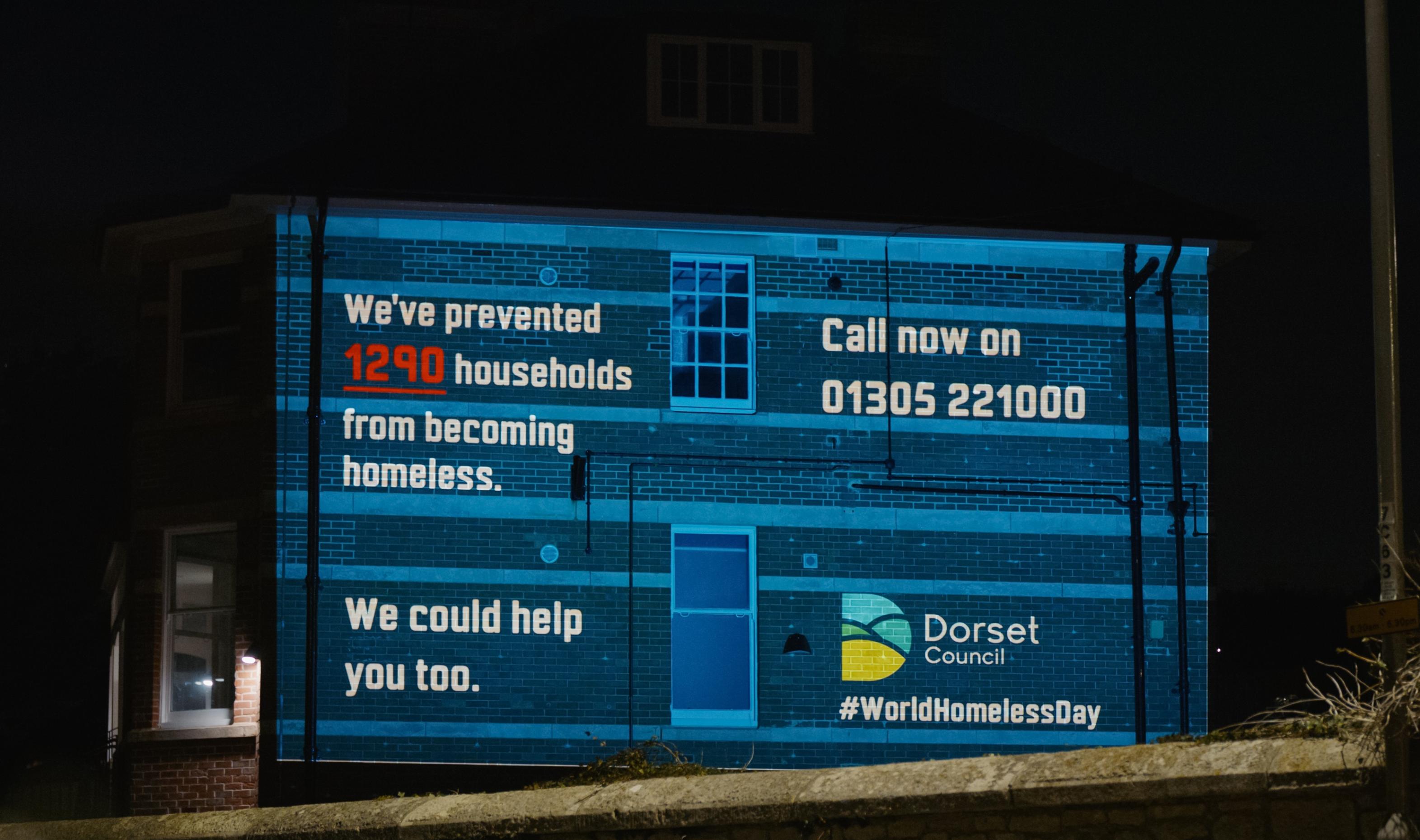 The side of a red brick building has been illuminated with a blue background with white text which has the council&#39;s housing helpline number on it with their logo and hash tag world homeless day