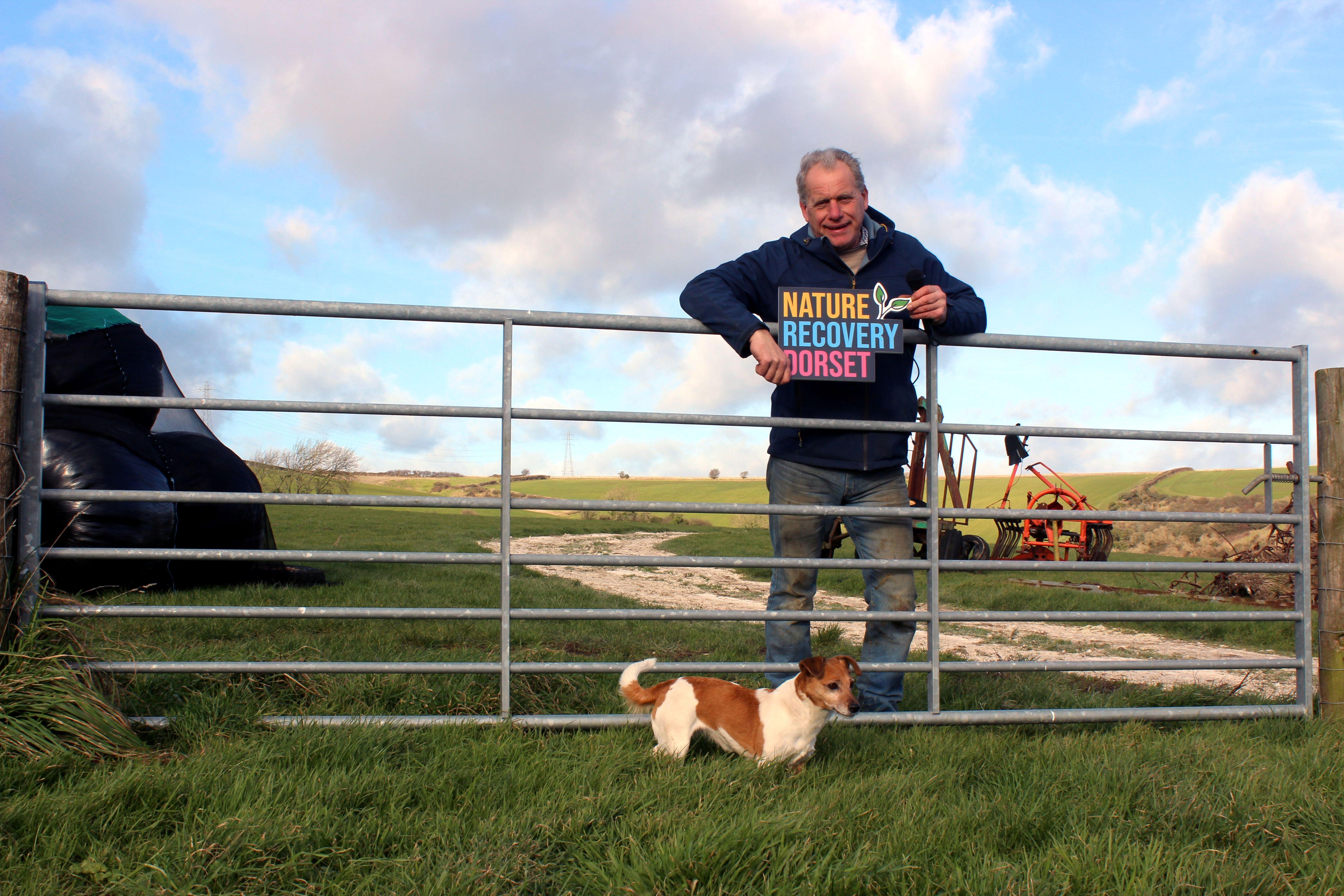 farmer stands behind farm gate holding a microphone and a Nature Recovery Dorset sign, there is a dog in front of the gate and you can see farmland behind the farmer