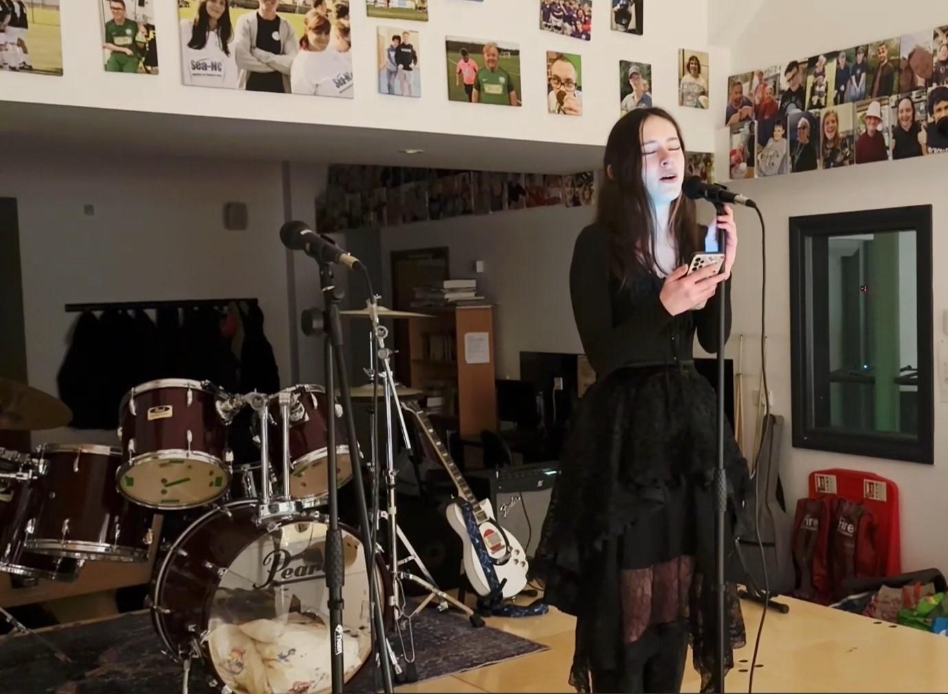 young person standing at a microphone holding a smartphone with drum set in the background