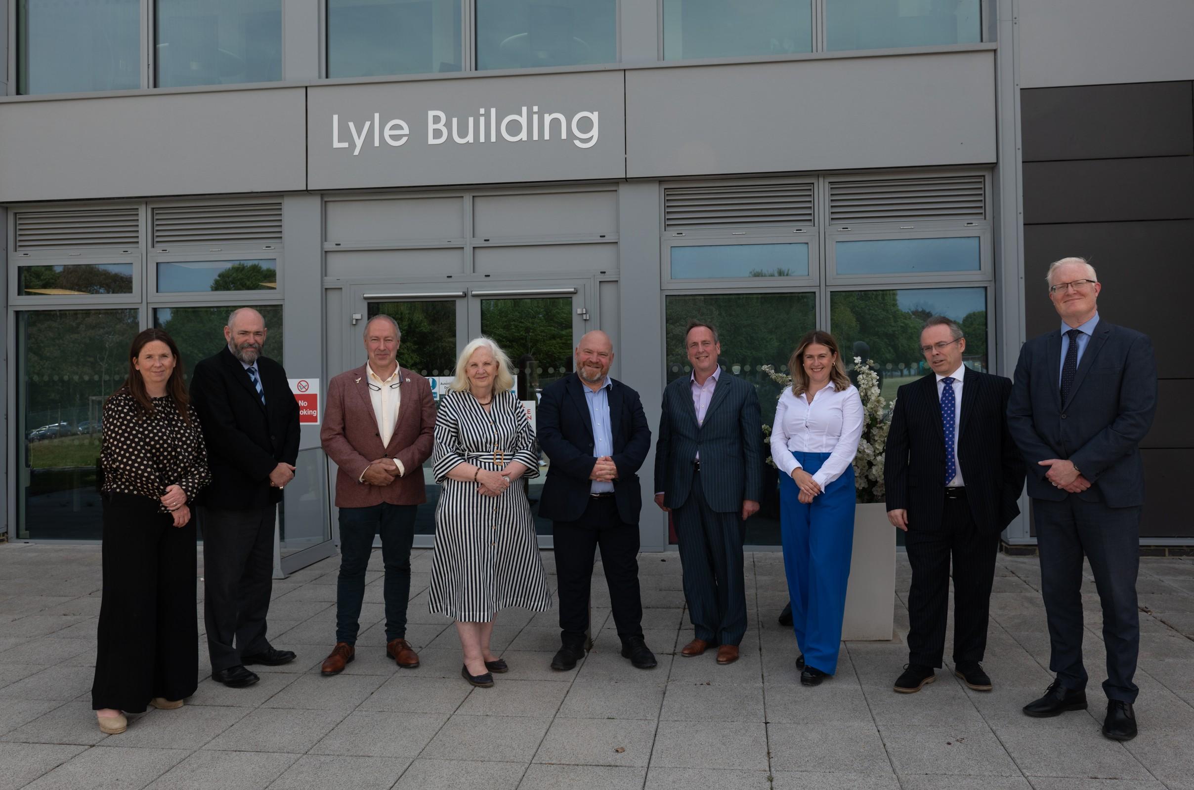 Nine men and women wearing smart clothes standing outside a grey, modern looking building