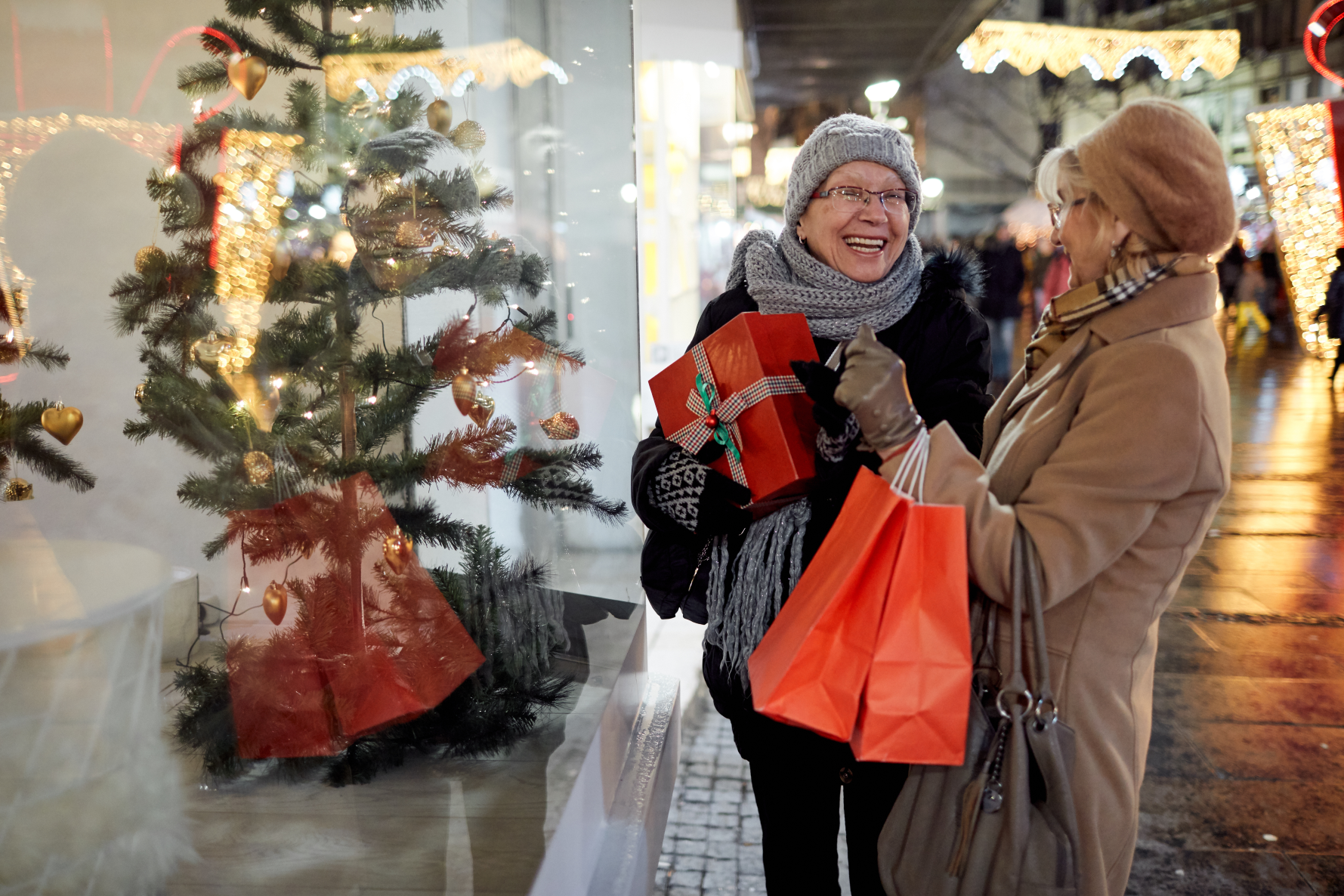 Two ladies holding shopping bags laughing in front of a shop window containing a Christmas tree