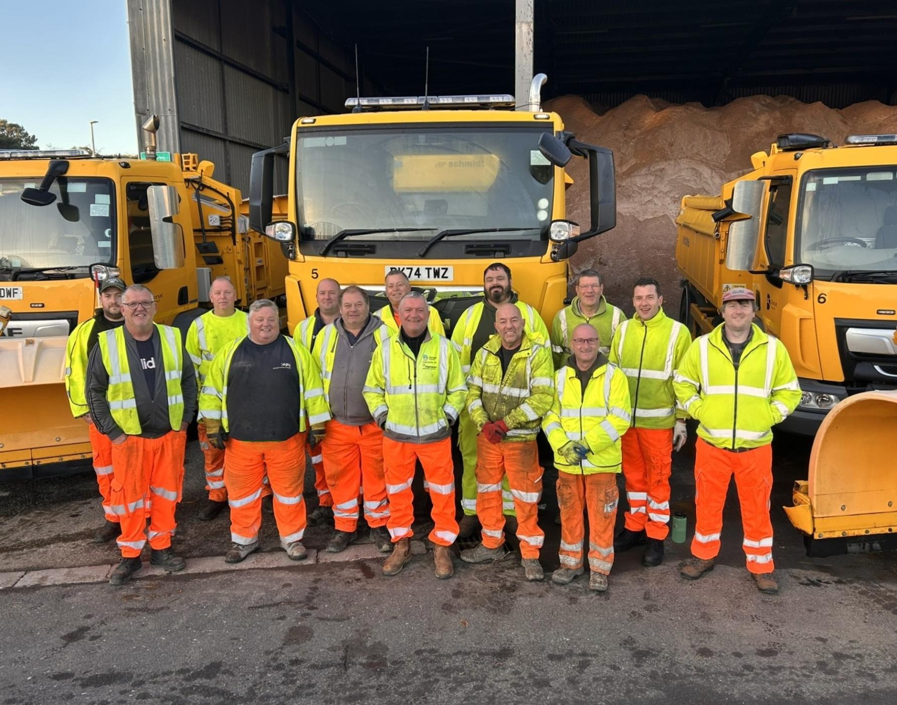 A line of up of some of the council&#39;s highways crew in front of some yellow gritting vehicles. They are wearing yellow jackets and orange trousers