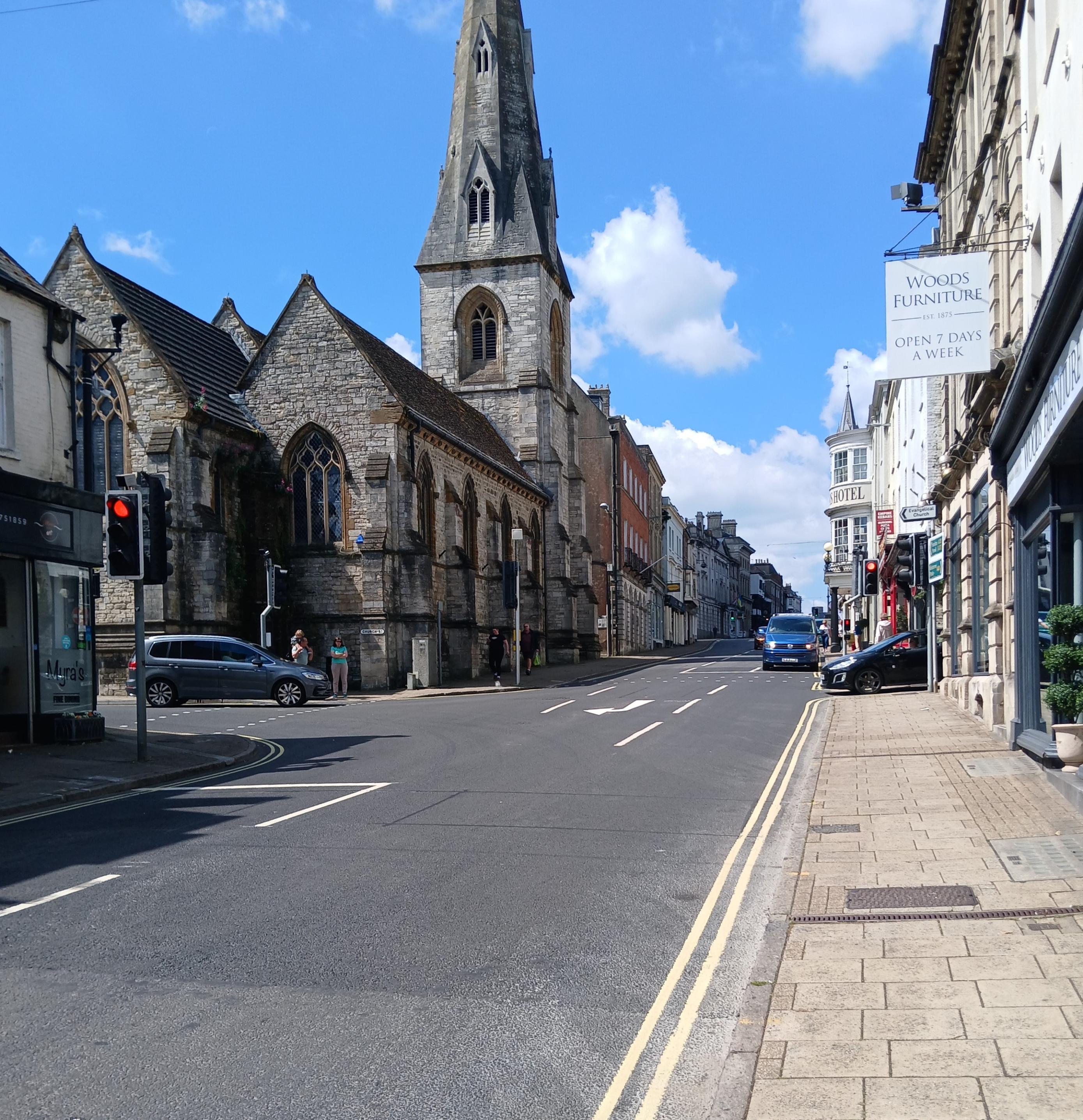 A main road on a sunny day with blue sky, a church in the background and cars waiting at junctions from two side roads. There is a red traffic light and a blue VW camper van waiting at another signal.
