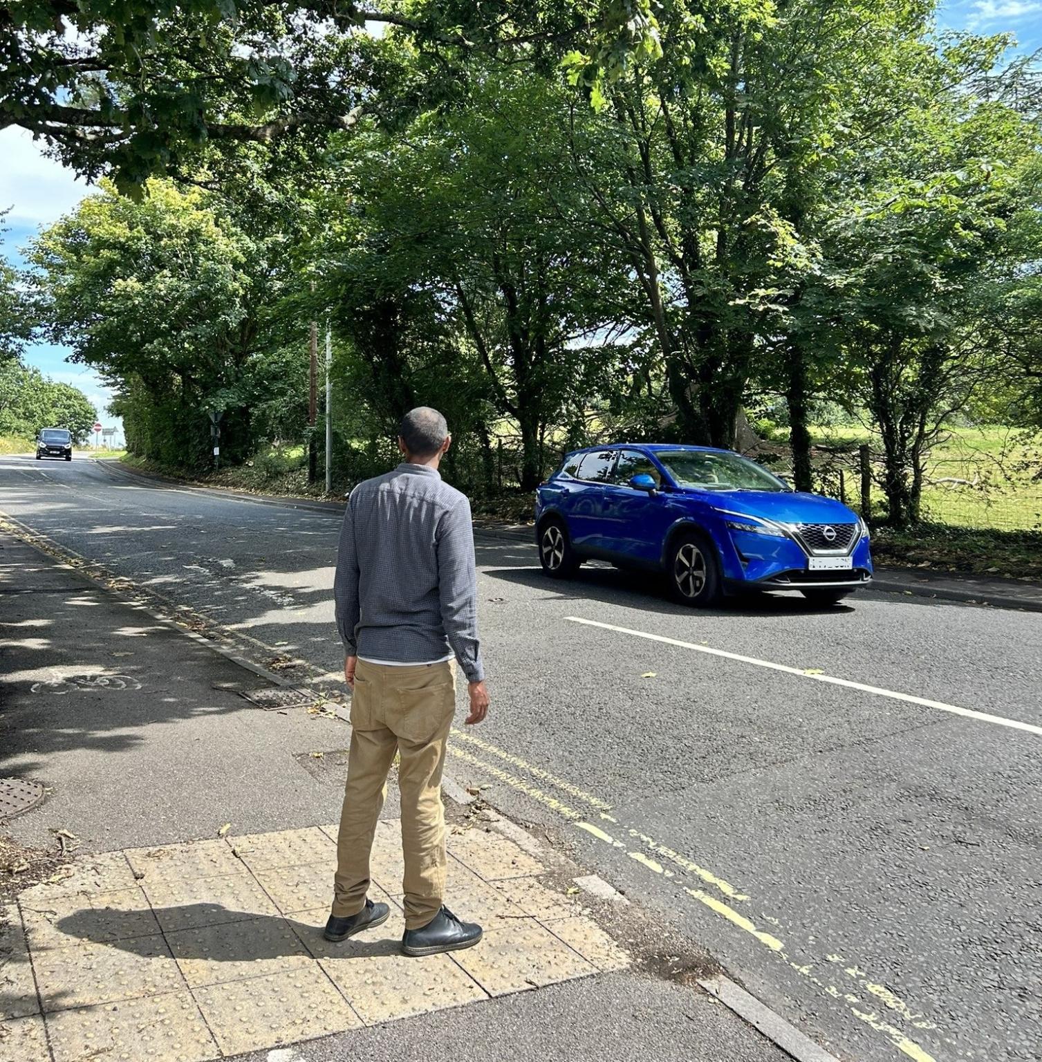 A person wearing a grey top and beige trousers waiting to cross a road on a sunny day where a blue car is passing