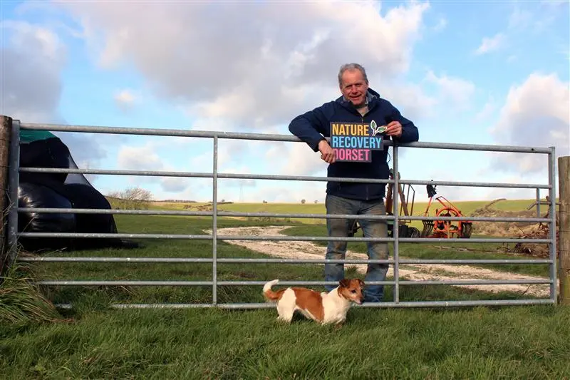 Person standing by a farm gate holding a “Nature Recovery Dorset” sign with a small dog in front.