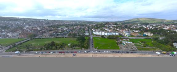 A road running parallel with a beach with green behind and further back there are buildings and hills and blue sky into the distance